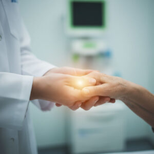 A close-up image of a healthcare professional's hands in a white lab coat gently holding the hand of an elderly patient. A soft, warm light emanates from between their clasped hands, with a blurred examination room in the background.